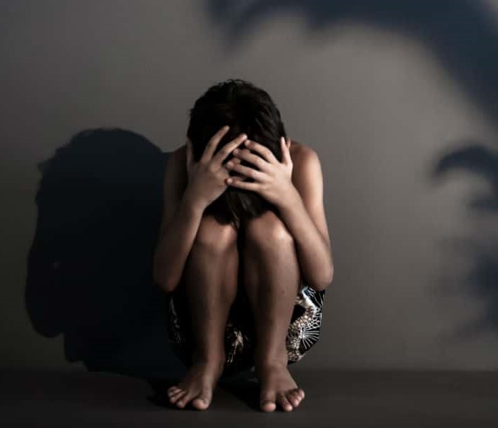 Foto von einem Mädchen, dass auf dem Boden sitzt die Hände über dem Kopf zusammengeschlagen hat weil sie Angst hat