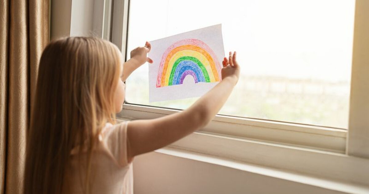 Foto von einem Mädchen das einen gezeichneten Regenbogen ans Fenster hält und lacht